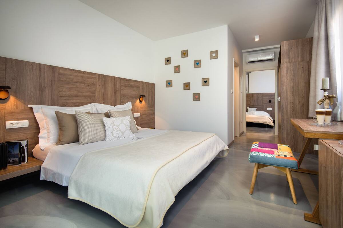 A modern bedroom with a wooden headboard, a bed with white bedding, a bedside table with a lamp, and a small stool.