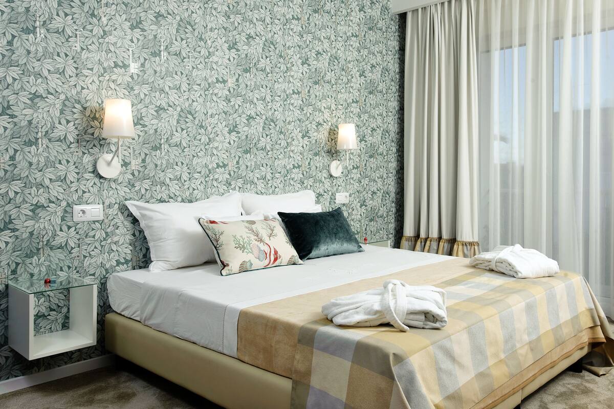 A neatly made bed with a checkered blanket, white pillows, and a dark throw pillow. There is a bedside table with a lamp and a power outlet. The room has a floral wallpaper and sheer curtains on the window.