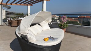 A wicker lounge chair with a white cushion and a magazine on it, a potted plant, and a view of buildings and mountains in the background. - Boban Luxury Suites (Split)