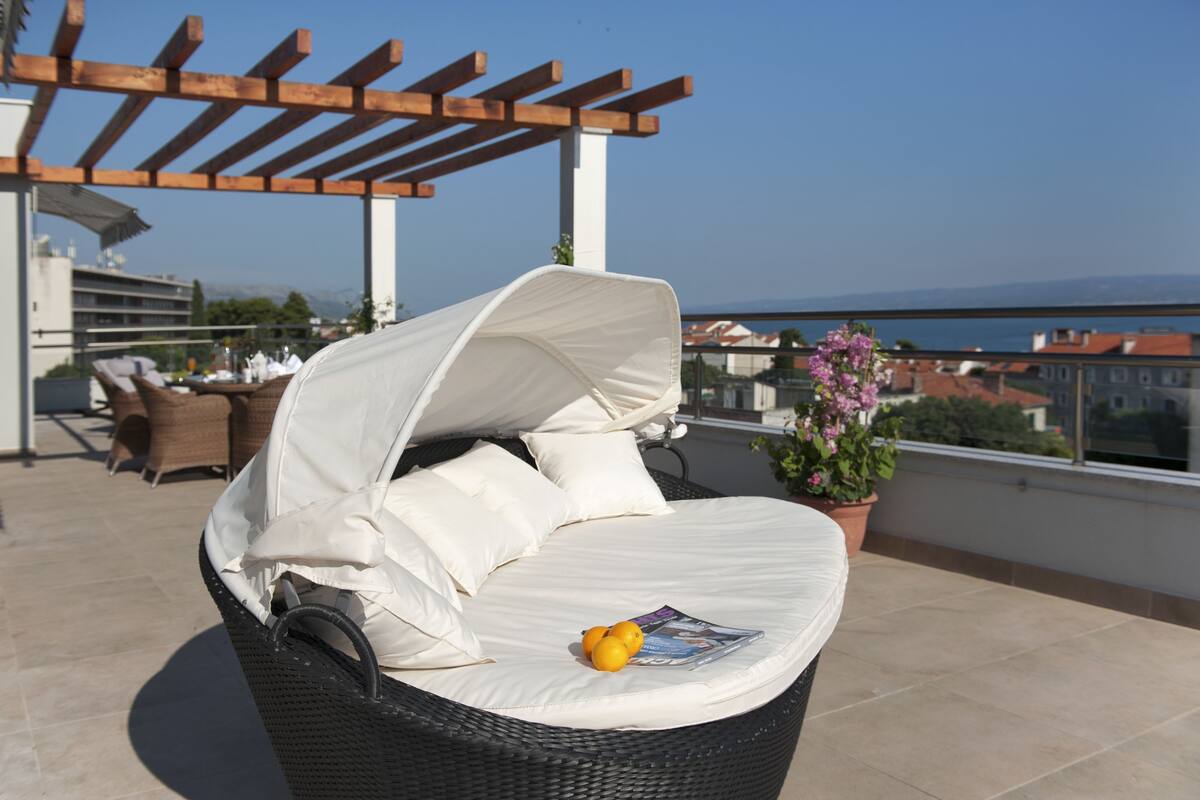 A wicker lounge chair with a white cushion and a magazine on it, a potted plant, and a view of buildings and mountains in the background.