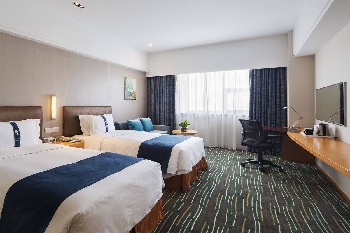 A hotel room with two beds, a desk with a computer, a chair, a small table with a plant, and a large window with curtains.