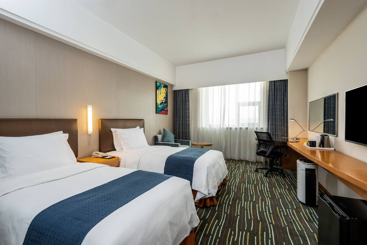 A hotel room with two beds, a desk, a chair, a TV, and a window with curtains.