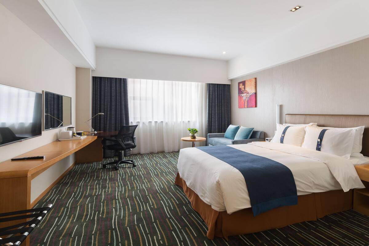A hotel room with a bed, a desk with a computer, a chair, a TV, and a window with curtains.