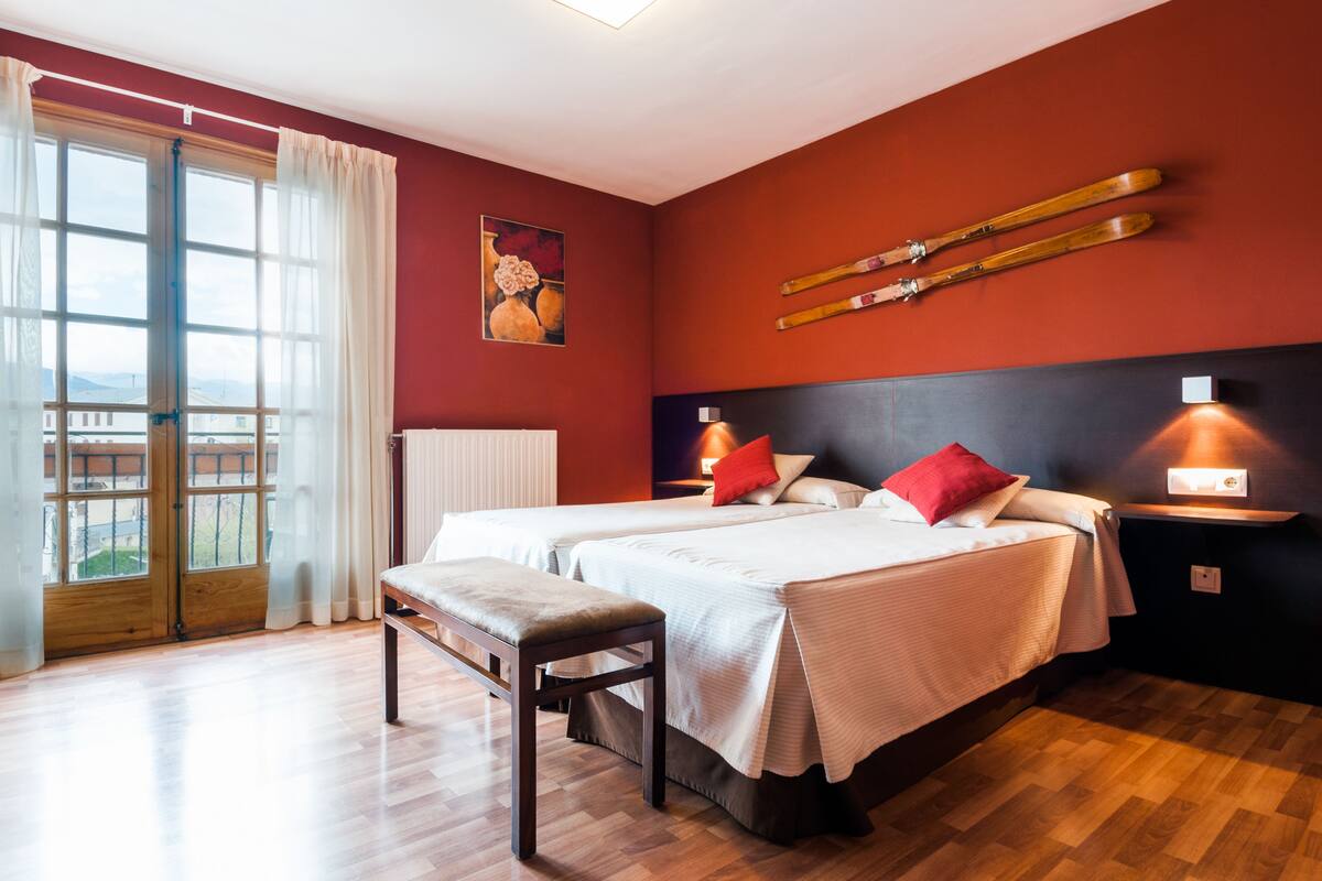 A hotel room with two beds, a bench, a view through a large window, and skis mounted on the wall.