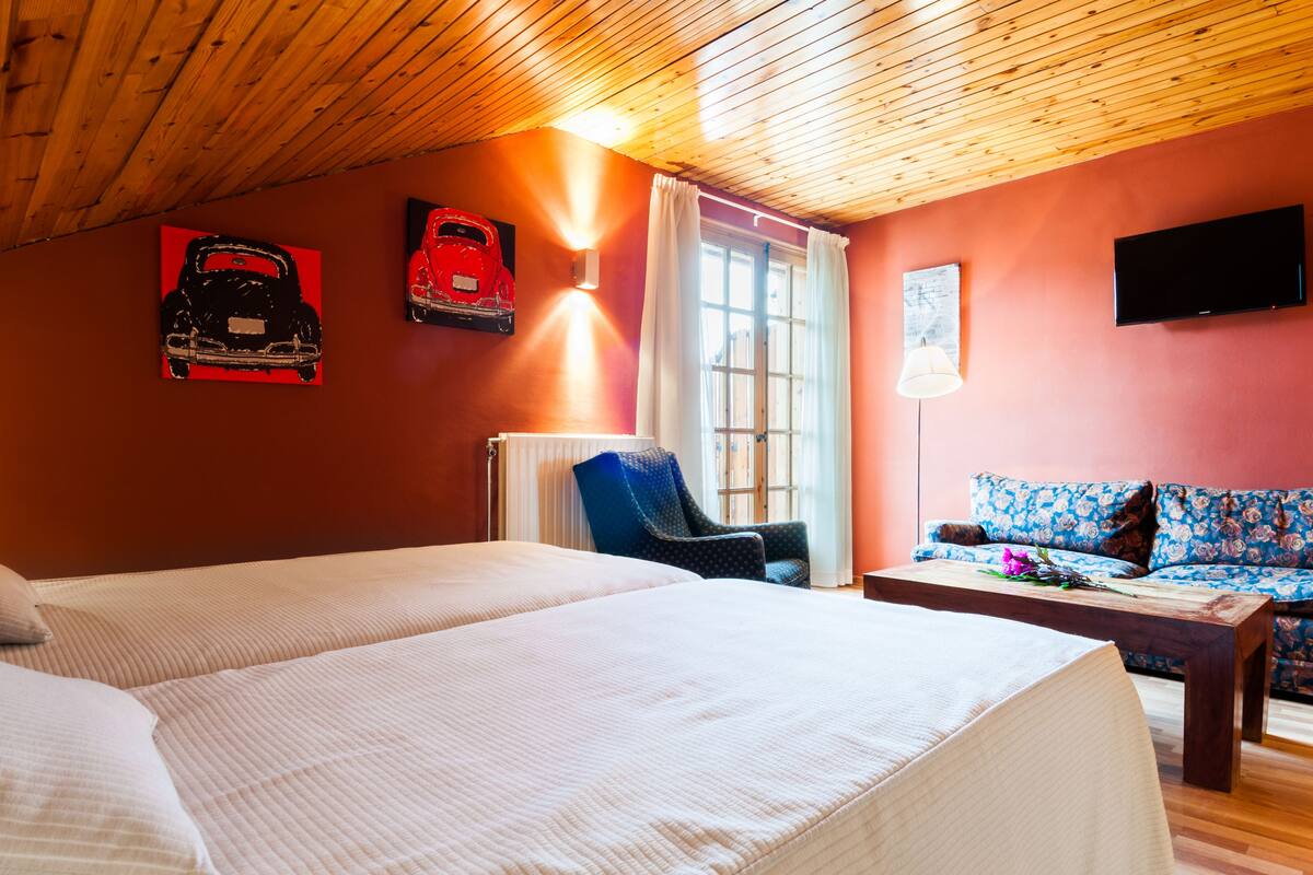 A room with two beds, a blue chair, a sofa, a table, a TV, and artwork on the walls.