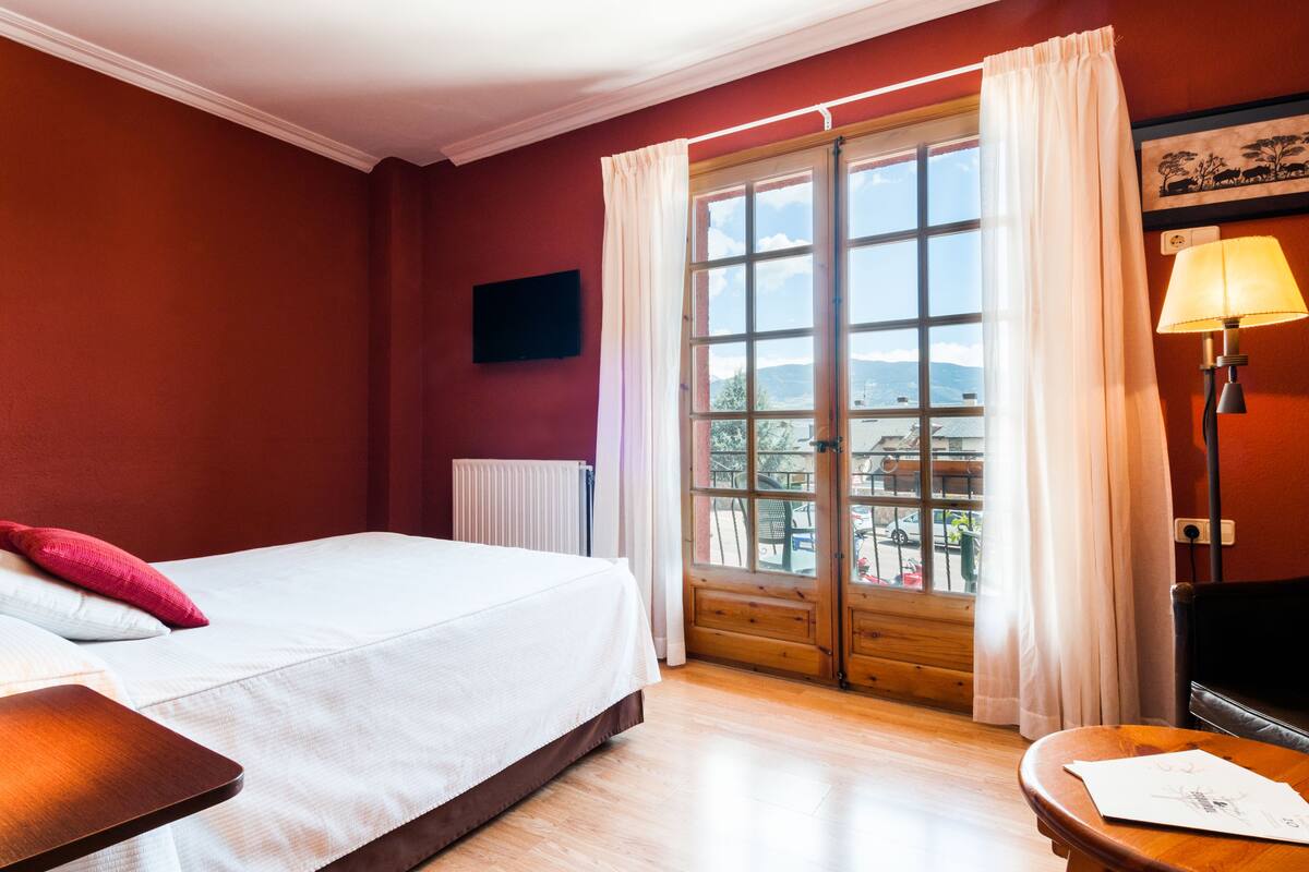 A hotel room with a bed, a wooden nightstand, a lamp, a window with curtains, and a view of the outside.