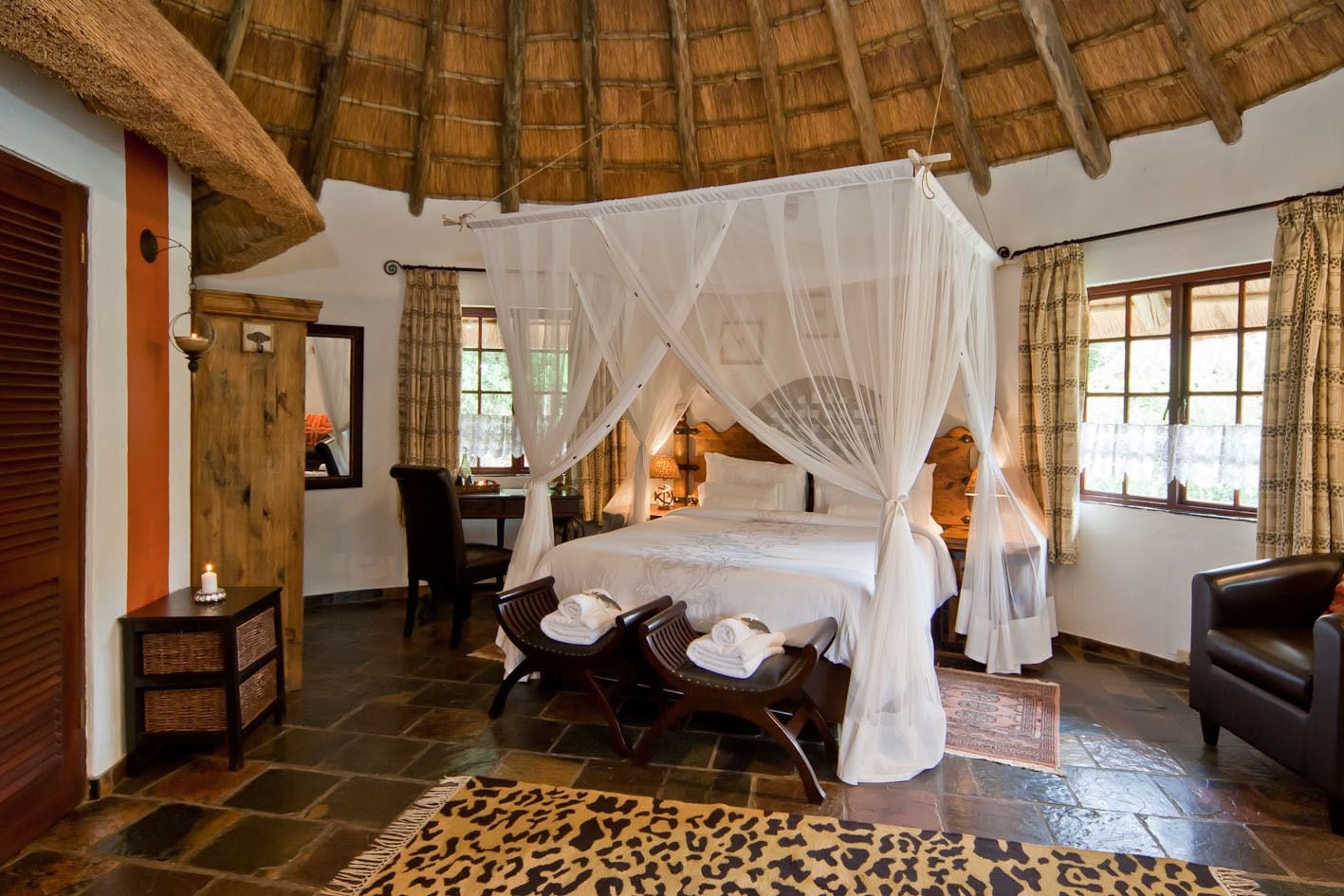 A room with a bed under a canopy, a desk with a chair, a mirror, and a window with curtains.