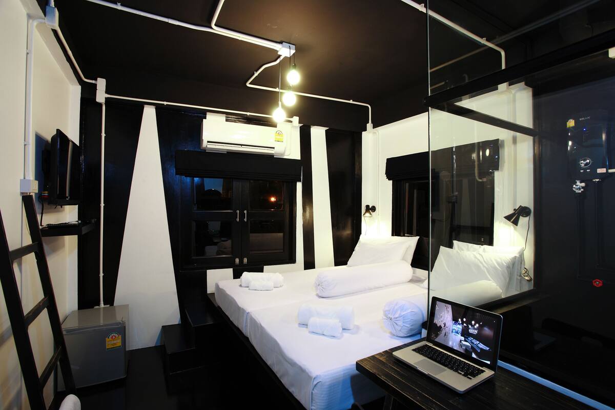 Una habitación de hotel moderna con cama, escritorio, laptop, un mueble pequeño y un espejo grande.