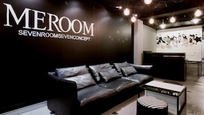 A modern interior with a black leather sofa, a white coffee table, and a wall displaying the text "MEROOM SEVENROOMSEVENCONCEPT". - Meroom (Phuket)