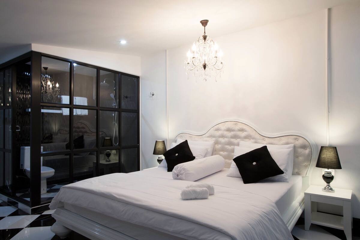 Un dormitorio moderno con una cama grande, dos mesitas de noche con lámparas, una araña y un baño con paredes de vidrio.