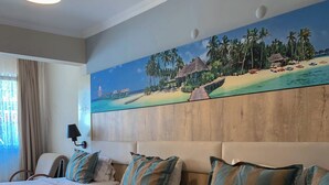 A hotel room with two beds, a large painting of a beach scene, a ceiling light, and a window with curtains. - Akkan Beach Hotel (Bodrum)
