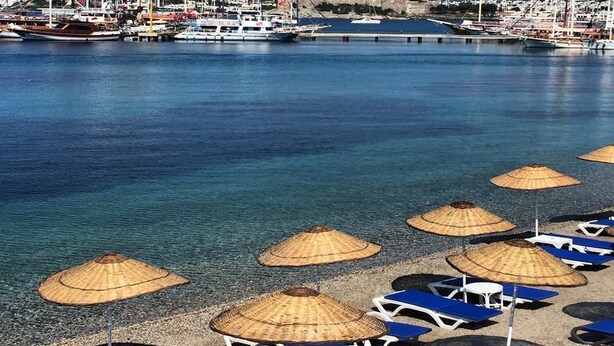 A beach with sun loungers and thatched umbrellas, a marina with boats, and a distant building. - Akkan Beach Hotel (Bodrum)