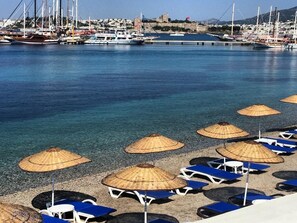 A beach with sun loungers and thatched umbrellas, a marina with boats, and a distant building. - Akkan Beach Hotel (Bodrum)