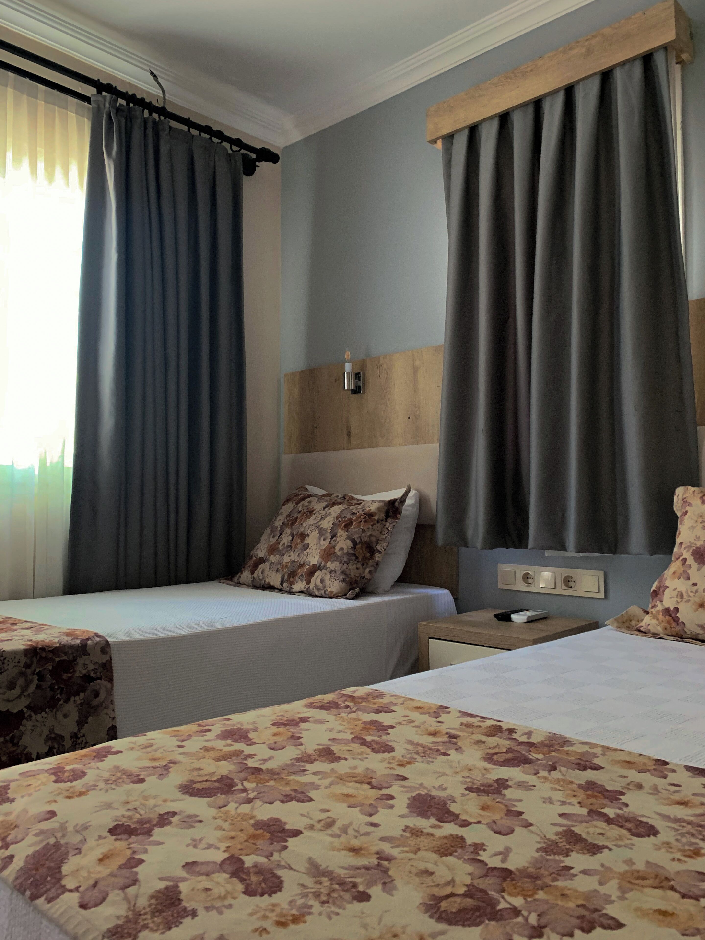 Habitación de hotel con dos camas, cabecero de madera, mesita de noche con control remoto y colcha estampada con motivos florales.
