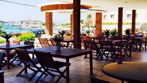 An outdoor dining area with tables and chairs, a view of the sea and a bridge, and a building in the background. - Akkan Beach Hotel (Bodrum)