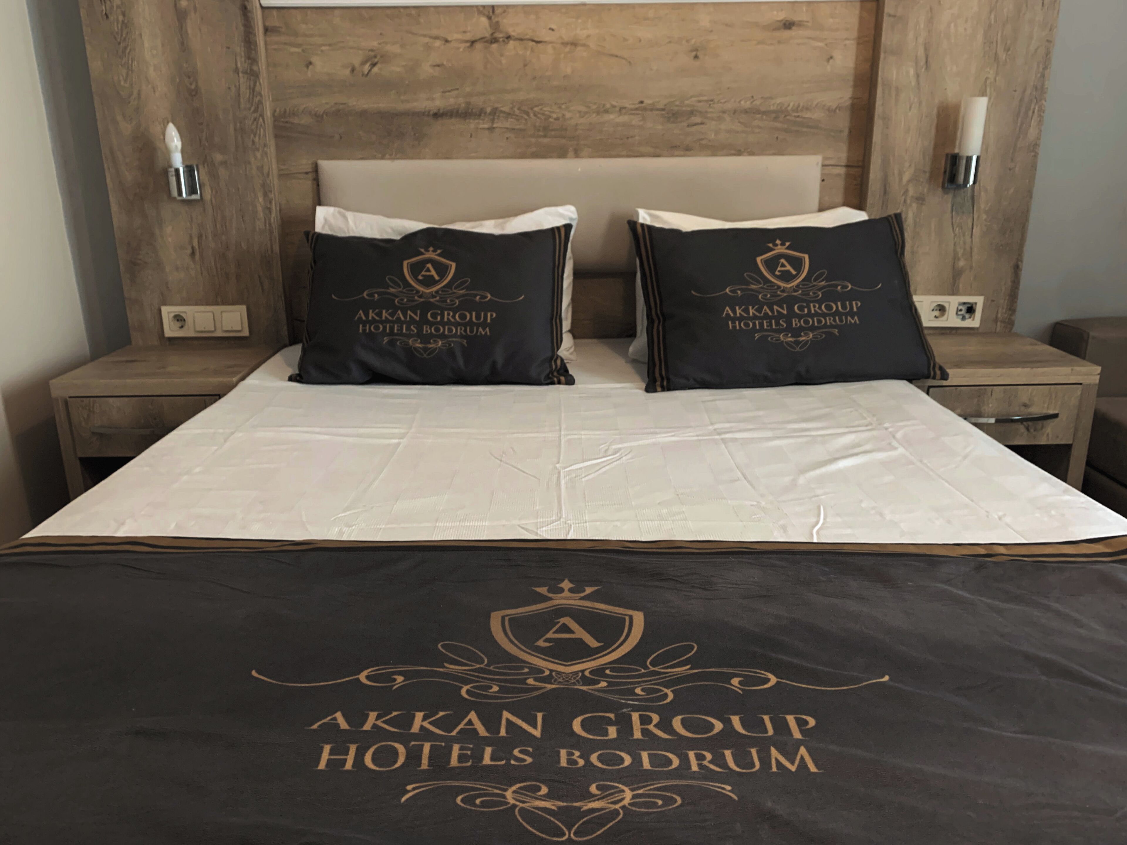 A hotel room with a bed, two pillows, a headboard, bedside tables, and a wall-mounted light fixture.