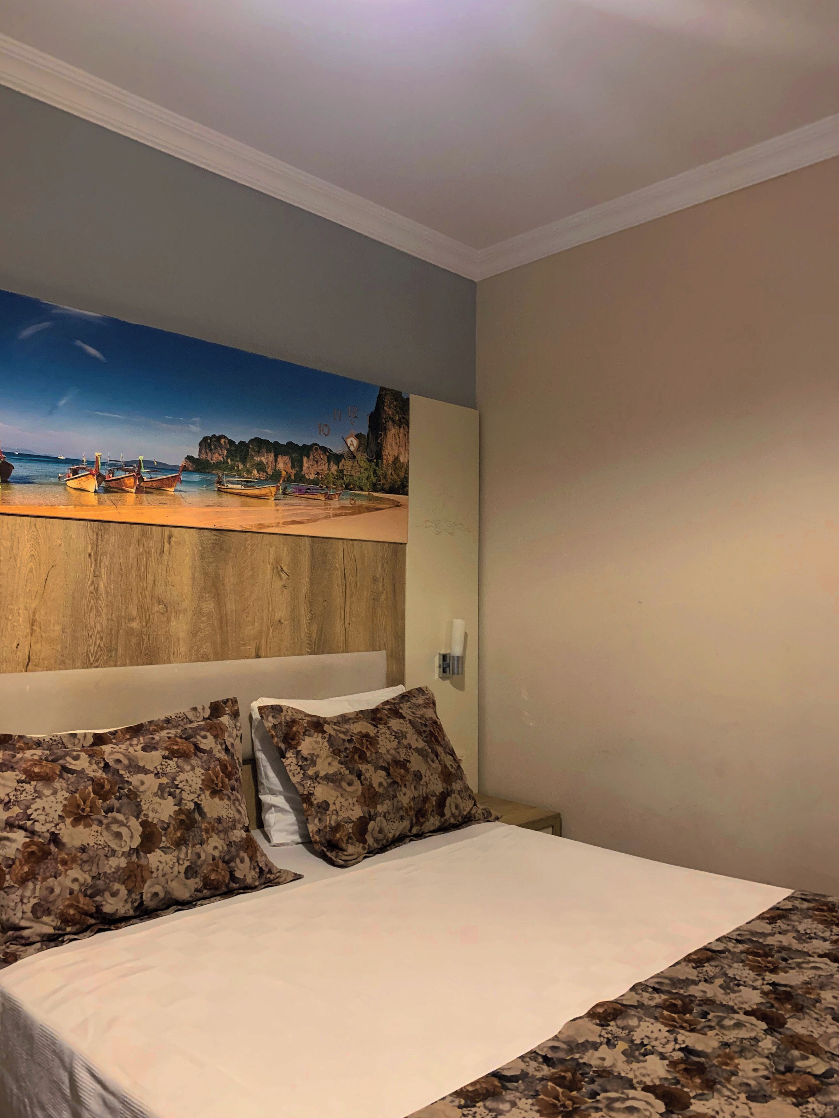A bedroom with a bed, floral pillows, a wooden headboard, a wall-mounted light, and a beach scene painting.