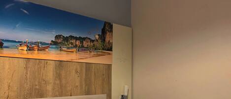 A bedroom with a bed, floral pillows, a wooden headboard, a wall-mounted light, and a beach scene painting.