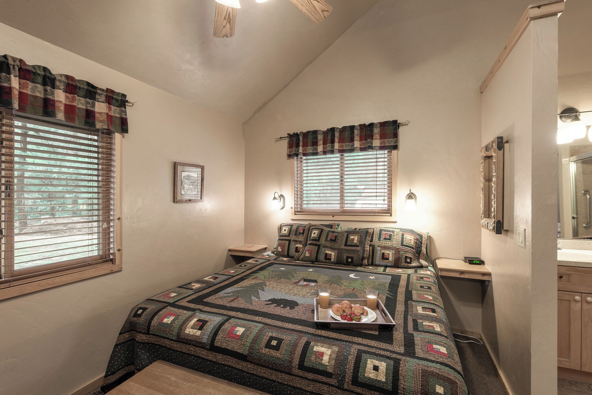 A bedroom with a large bed, two bedside tables, a nightstand, a window with a plaid valance, and a bathroom visible to the right.