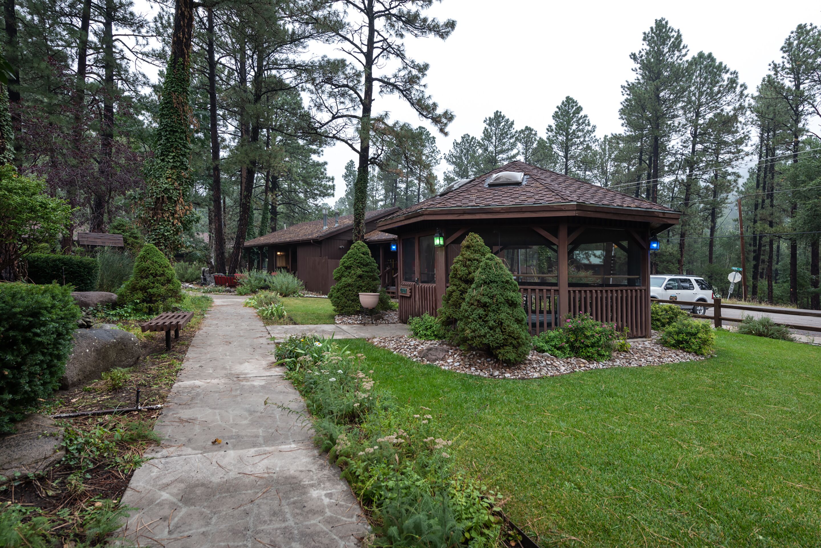 Eine Lodge mit einem Kiesweg, der zu einem hölzernen Gazebo führt, umgeben von einem gepflegten Garten und hohen Kiefern.