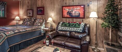 A cozy room with a wooden wall, a brown leather couch, a bed with a quilt, and a small table with a bottle and glasses.