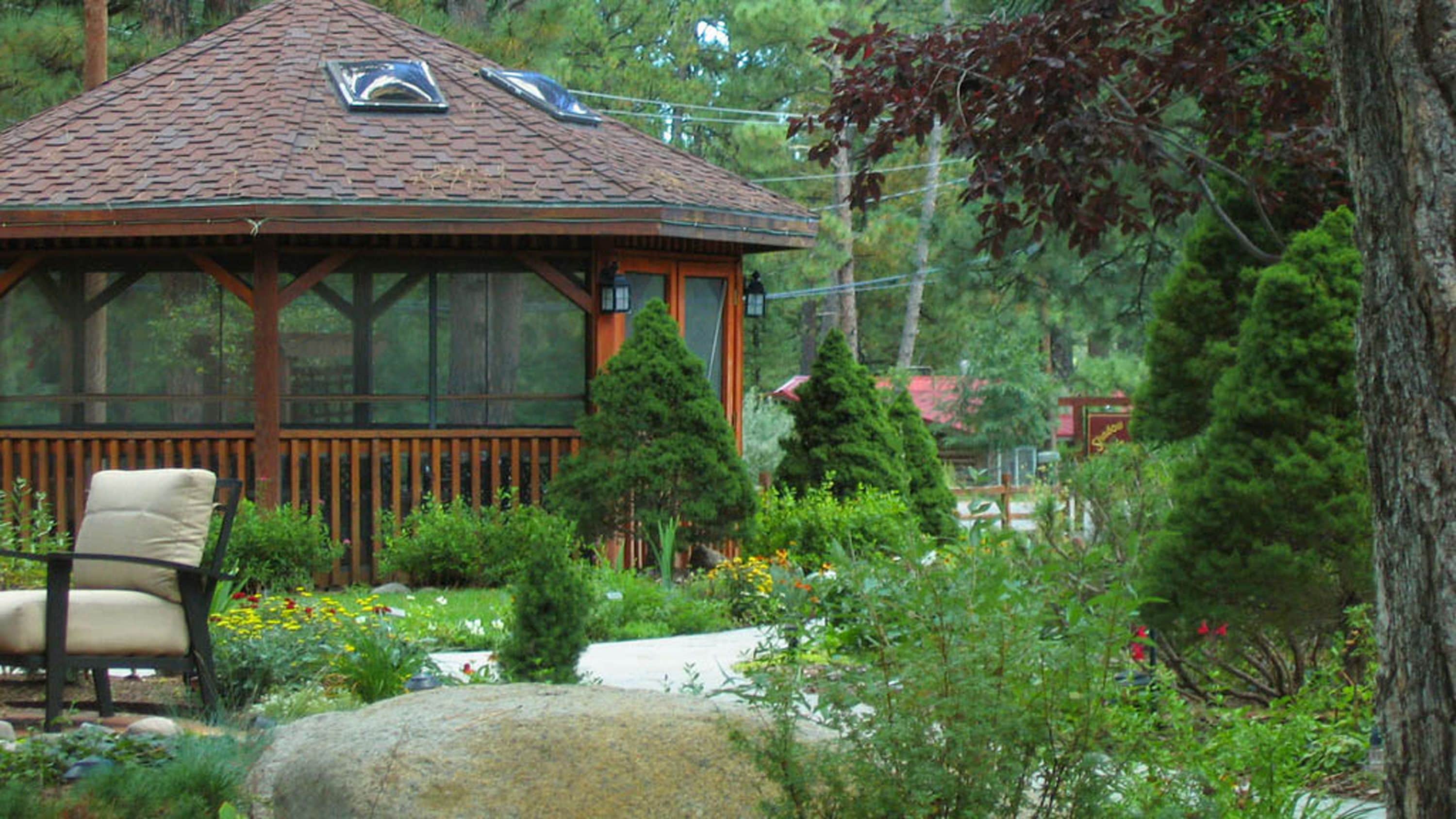 A gazebo with a tiled roof and large windows, surrounded by lush greenery and a stone bench.