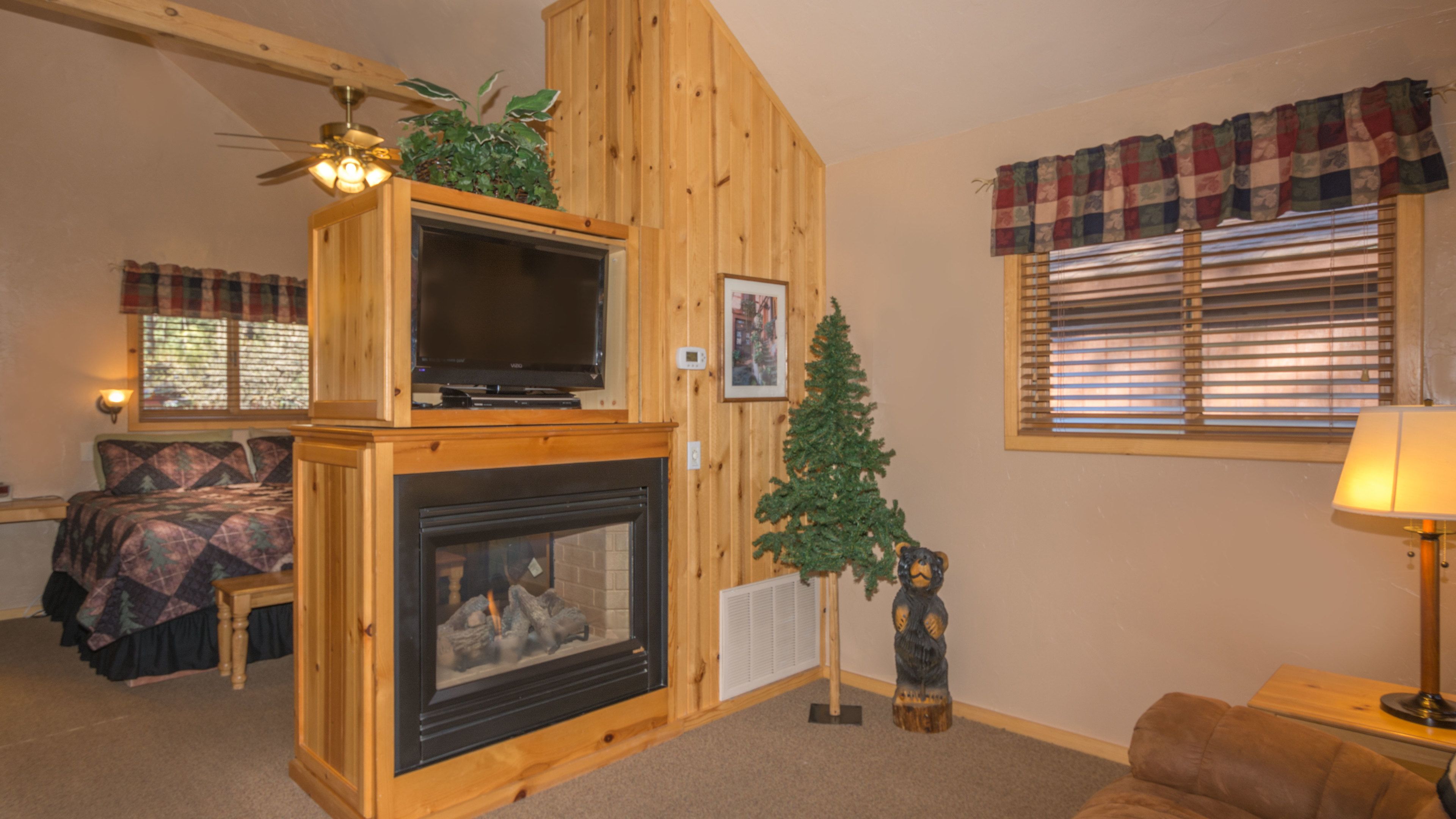 A cozy living room with a fireplace, wooden walls, a ceiling fan, a television, a bed, and a Christmas tree.