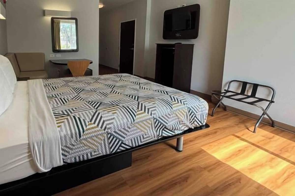 A hotel room with a bed, a desk, a chair, a TV, and a bench.