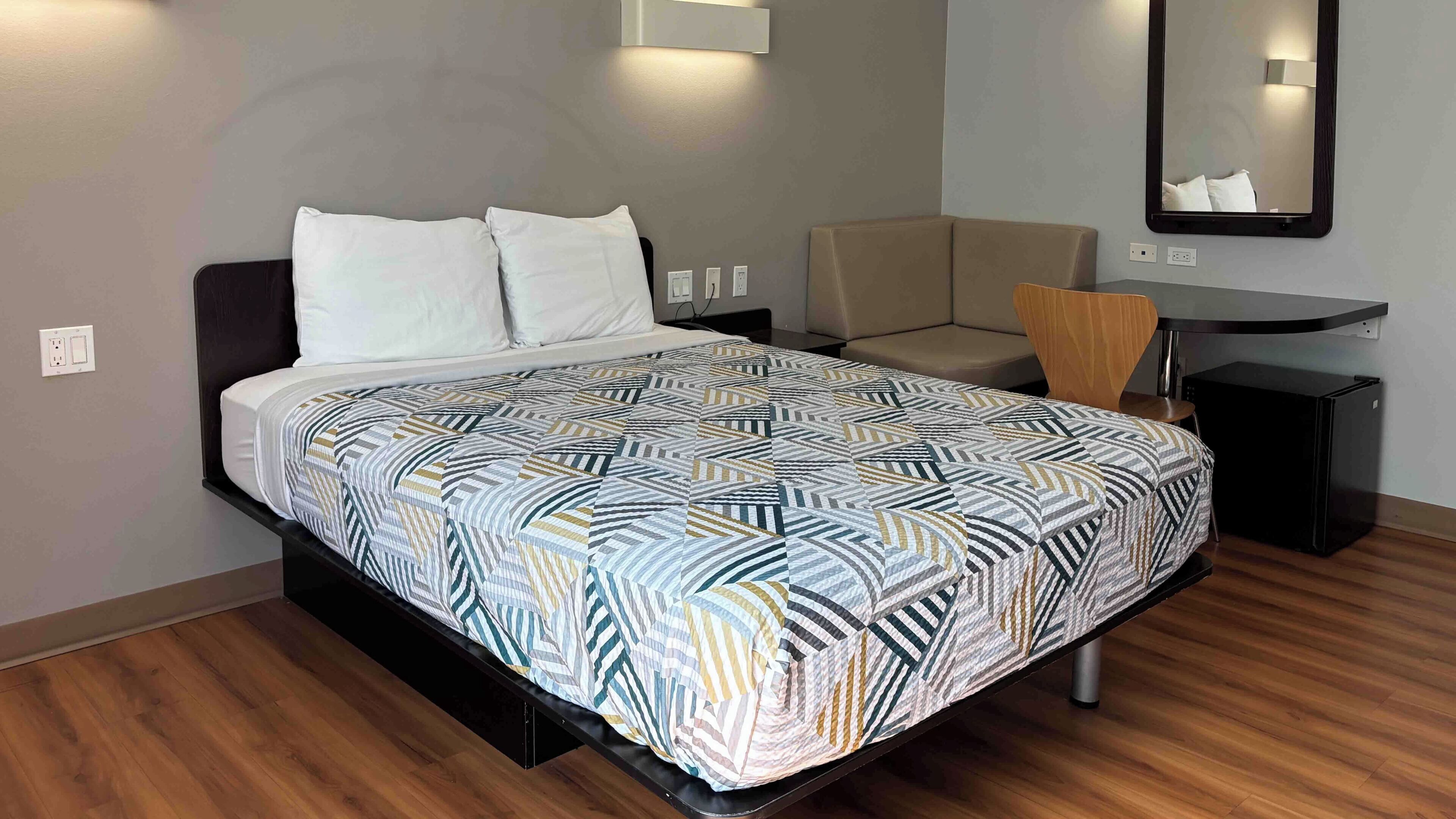 A modern hotel room with a bed, a desk, a chair, and a mirror.