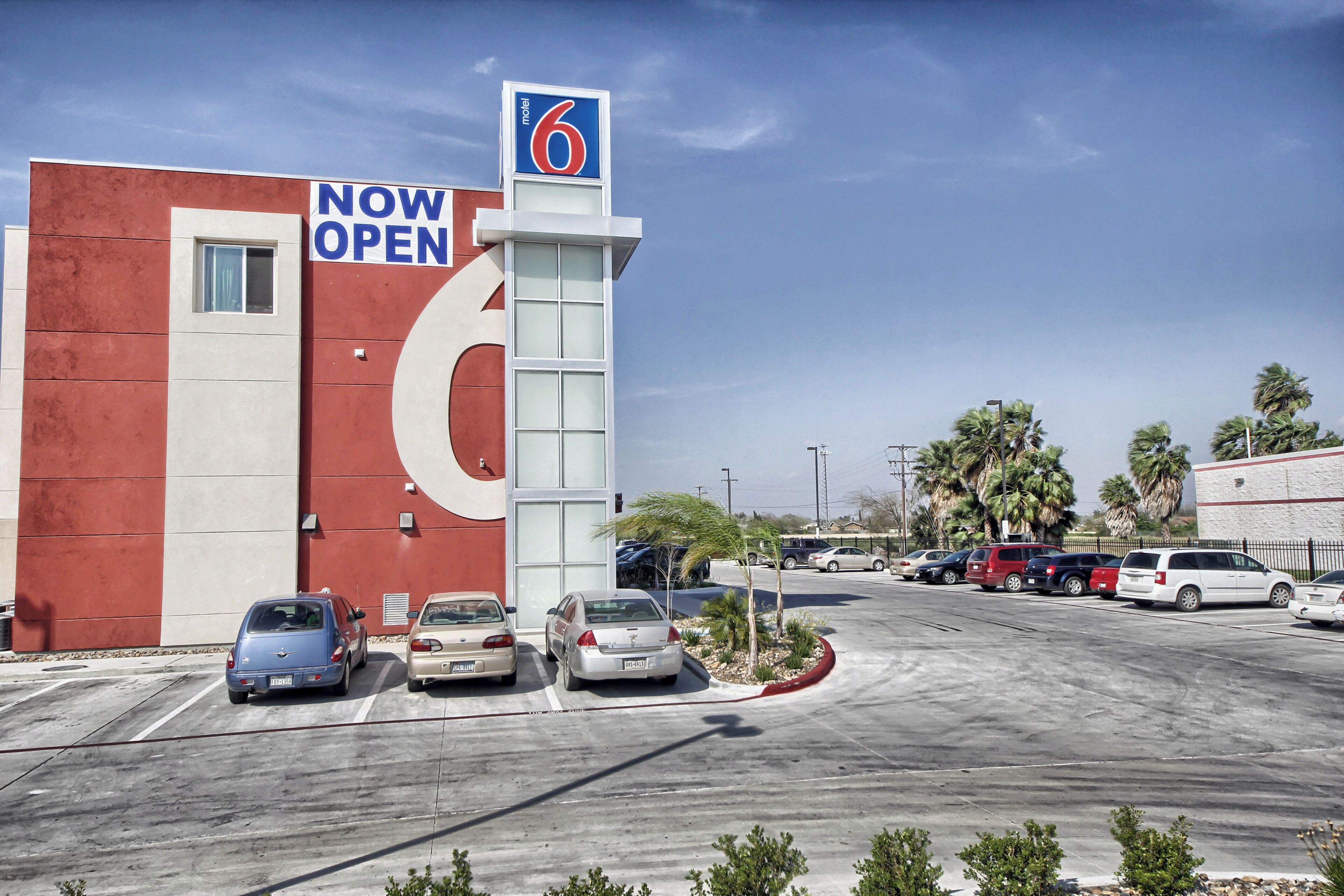 A motel with a 'NOW OPEN' sign, a large number '6' on the building, and several parked cars in the parking lot.