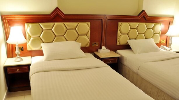 Ein Hotelzimmer mit zwei Einzelbetten, jeweils mit einem Kopfende mit geometrischem Muster. Es gibt Nachttische mit Lampen und eine kleine Schublade.
