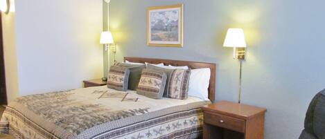 Una cama bien hecha con ropa de cama estampada, dos lámparas de noche, un cuadro en la pared y una silla en la esquina.