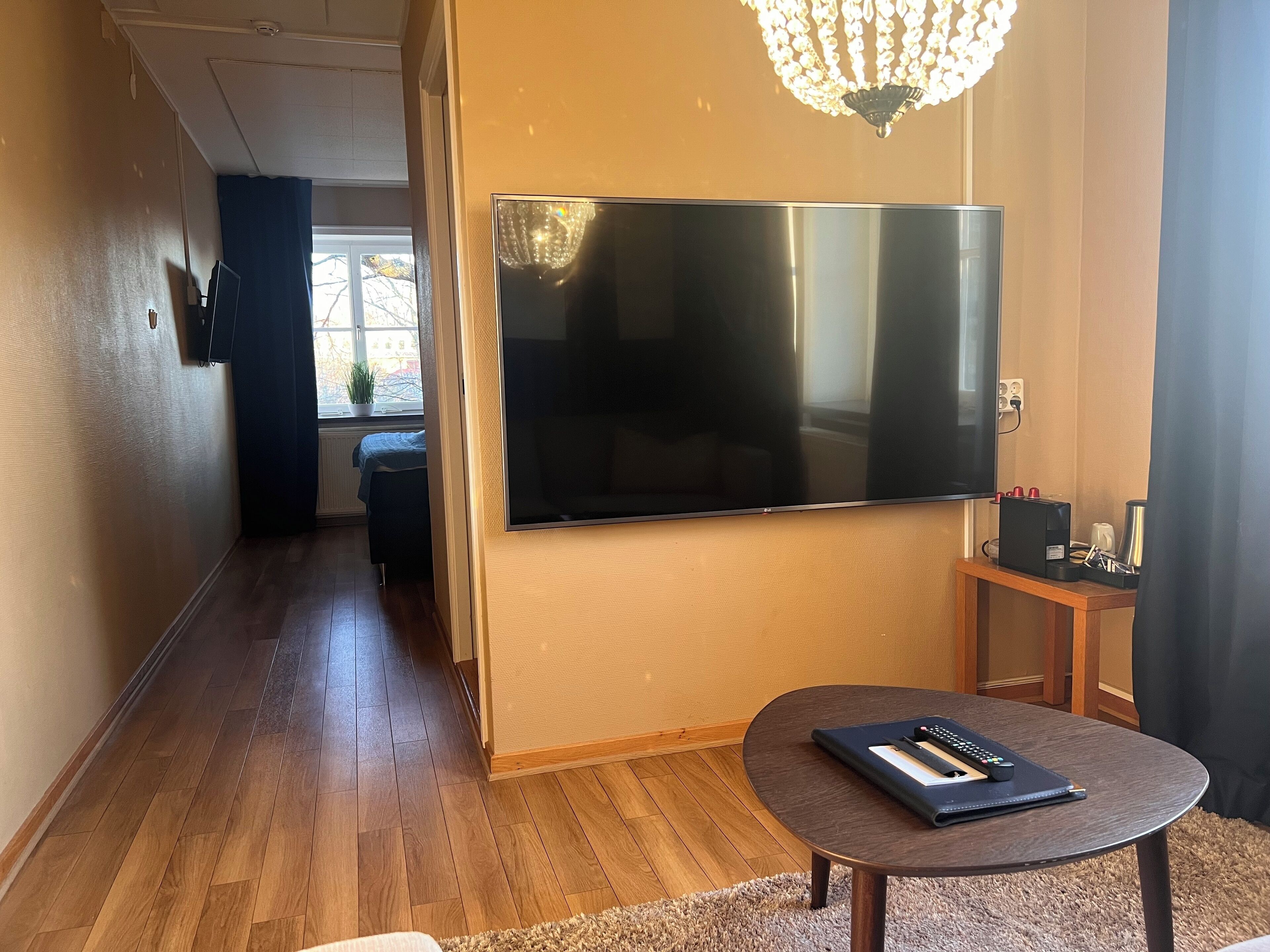 Una habitación moderna con un televisor de pantalla plana grande montado en la pared, una pequeña mesa de madera con un control remoto y una cafetera sobre una mesita auxiliar.
