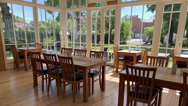 A spacious indoor dining area with wooden tables and chairs, large windows offering views of greenery and buildings. - Hotel Wictoria (Mariestad)