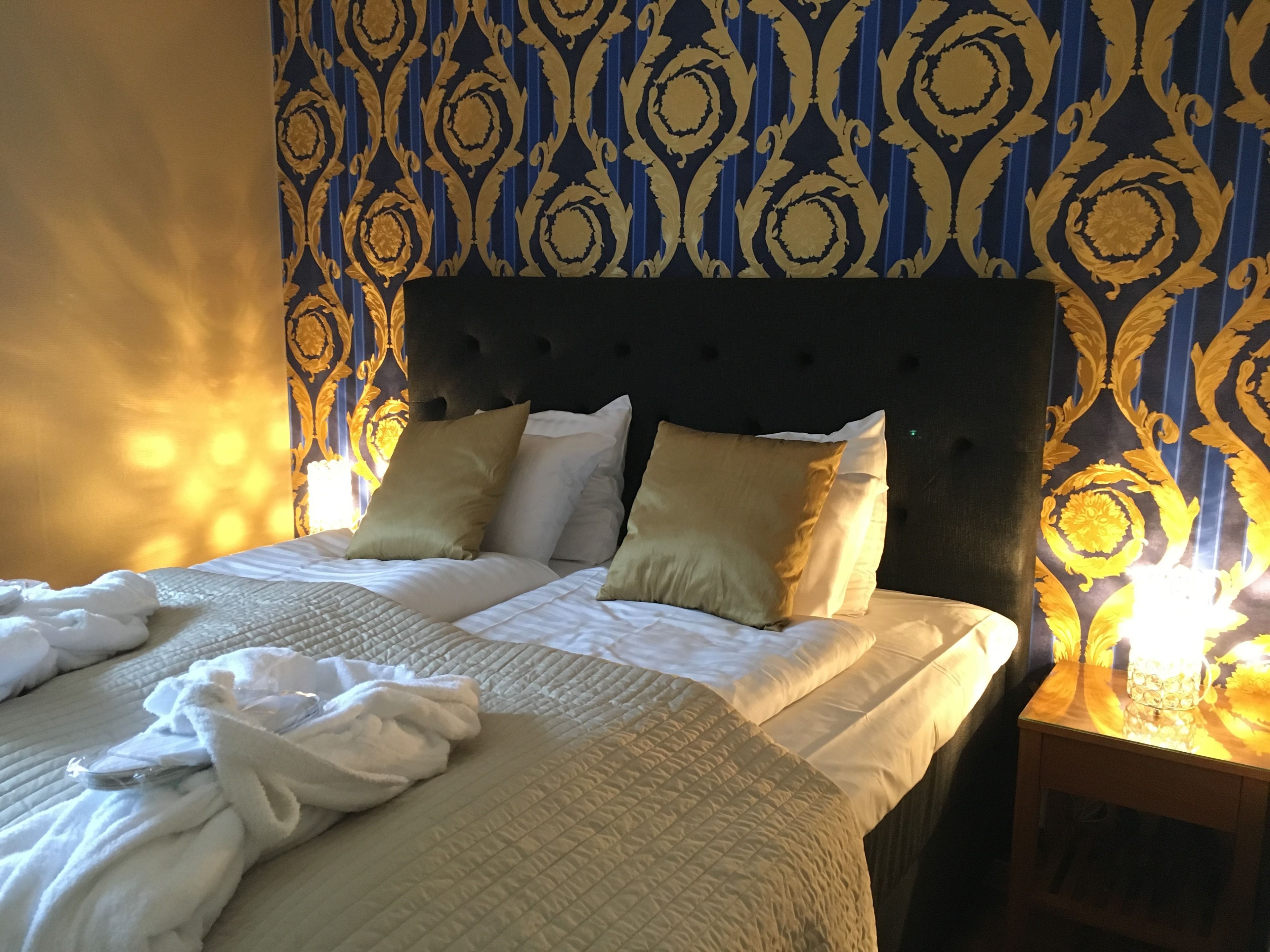 A neatly made bed with white linens and a beige quilt, a dark headboard, and a patterned wallpaper in blue and gold.