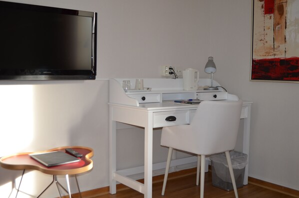 Un escritorio blanco con una silla, un televisor de pantalla plana montado en la pared, una mesita con una computadora portátil y un cesto de basura.