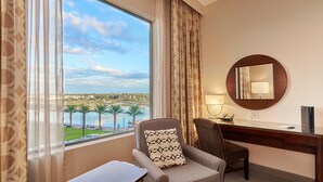 Ein Hotelzimmer mit großem Fenster, das Blick auf ein Gewässer und Palmen bietet, einem Schreibtisch mit Stuhl und einem runden Spiegel an der Wand.