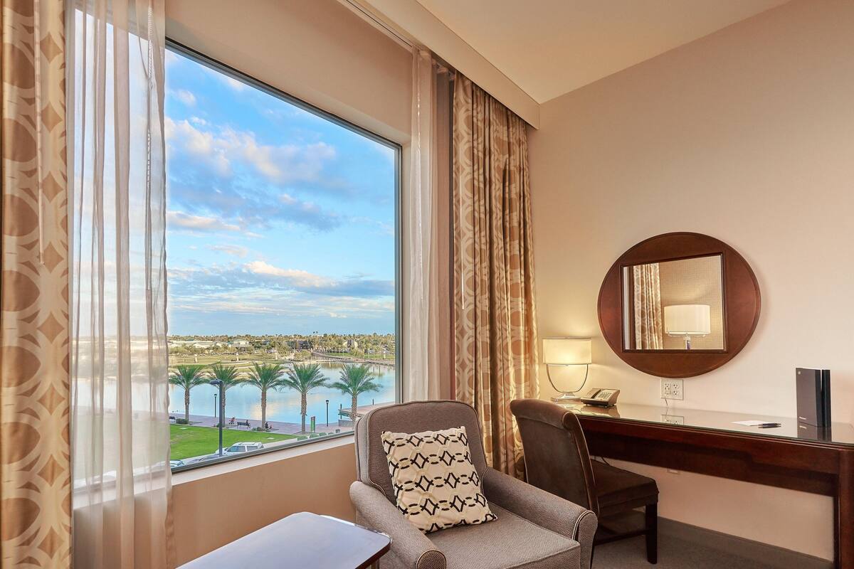 A hotel room with a large window offering a view of a body of water and palm trees, a desk with a chair, and a round mirror on the wall.