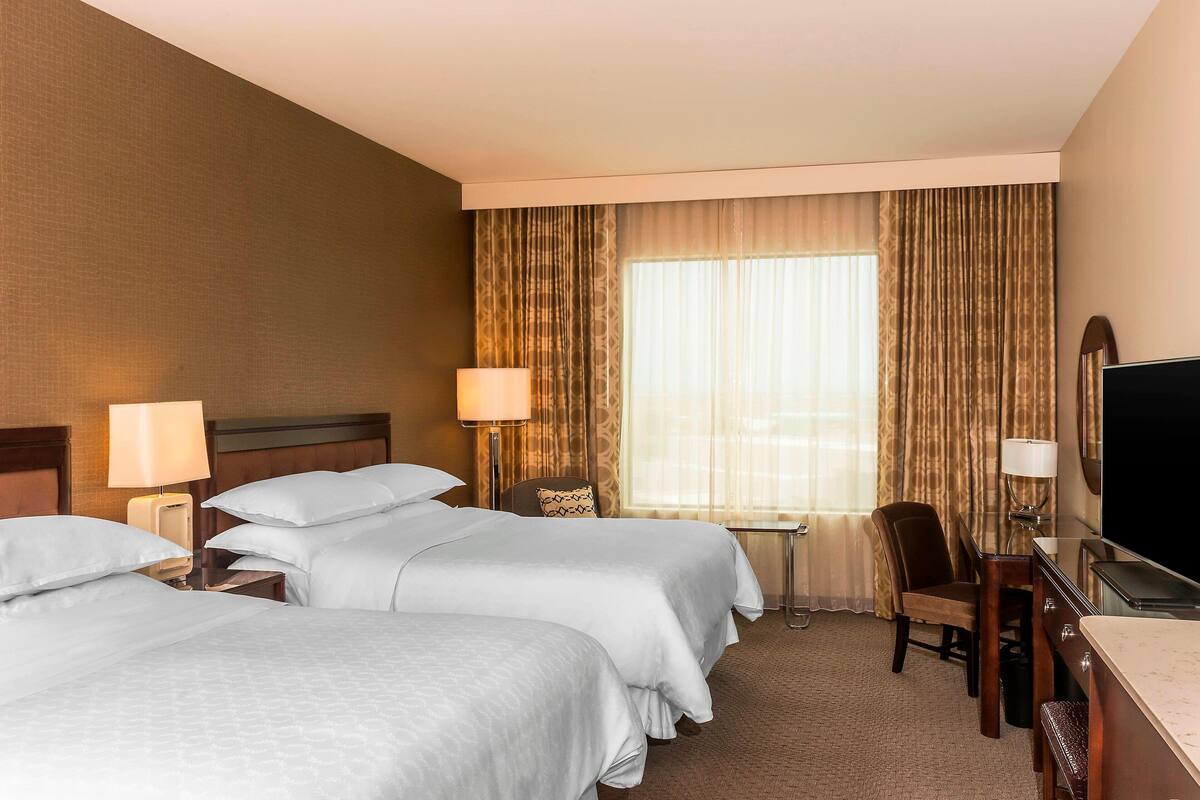 A hotel room with two beds, a desk, a chair, a TV, and a window with curtains.