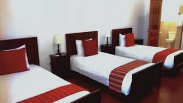 Habitación de hotel con tres camas individuales, cada una con almohada roja y colcha blanca. Cada cama tiene un cabecero de madera y una mesita de noche con lámpara.