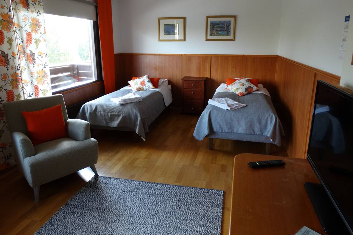 A hotel room with two single beds, a wooden headboard, a small desk with a TV, a chair, and a window with curtains.