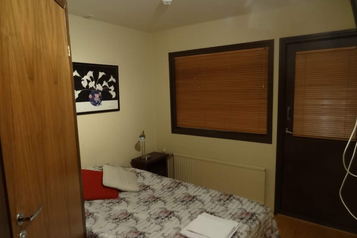 A bedroom with a bed, a nightstand, a wall-mounted lamp, a window with blinds, and a door.