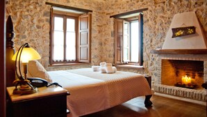 A bedroom with stone walls, a fireplace, a bed with a quilt, and a nightstand with a lamp and phone. - Archontiko Hatzipanayiotis (South Kynouria)