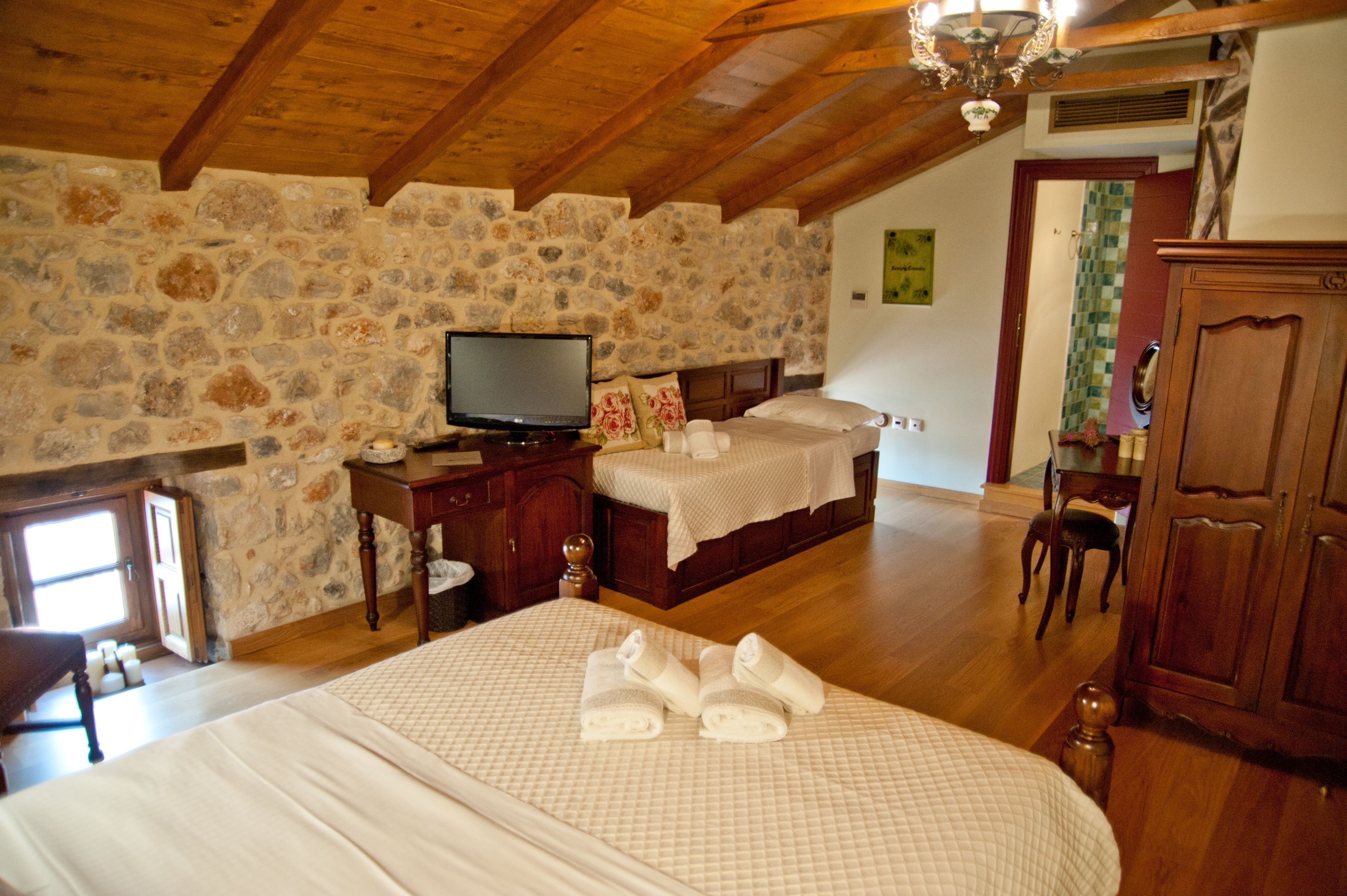 Un dormitorio con pared de piedra, vigas de madera en el techo, una cama con colcha acolchada, un televisor sobre un aparador de madera y un armario de madera.