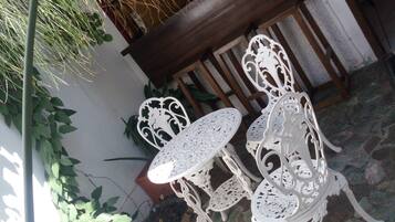 An outdoor patio with white wrought iron tables and chairs, surrounded by greenery and a stone floor.
