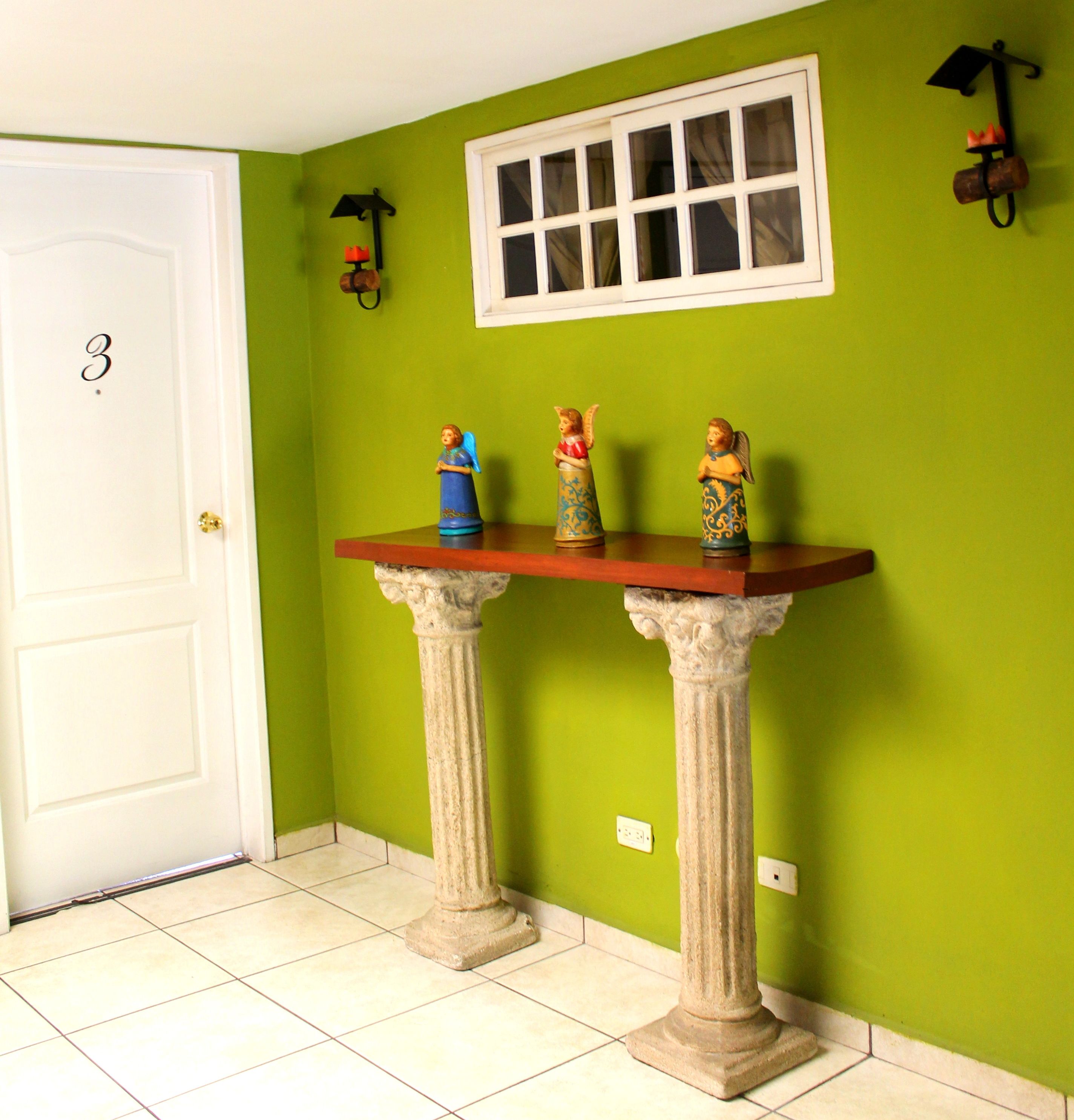 A hallway with a green wall, a white door marked with the number 3, a window with white frames, and a small table with three angel figurines.
