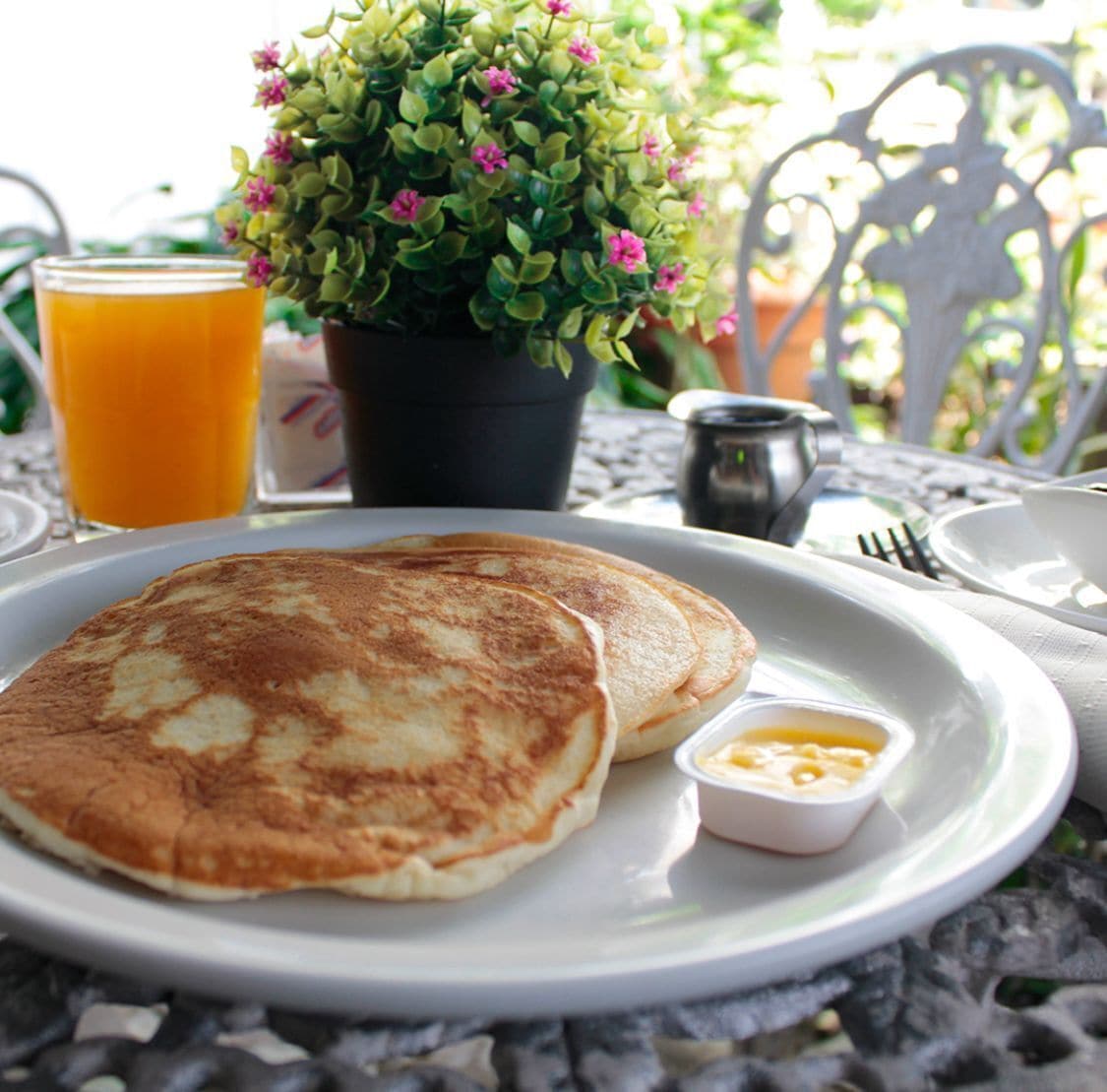 Een stapel pannenkoeken met een klein kommetje siroop, een glas sinaasappelsap en een potplant op een tafel.