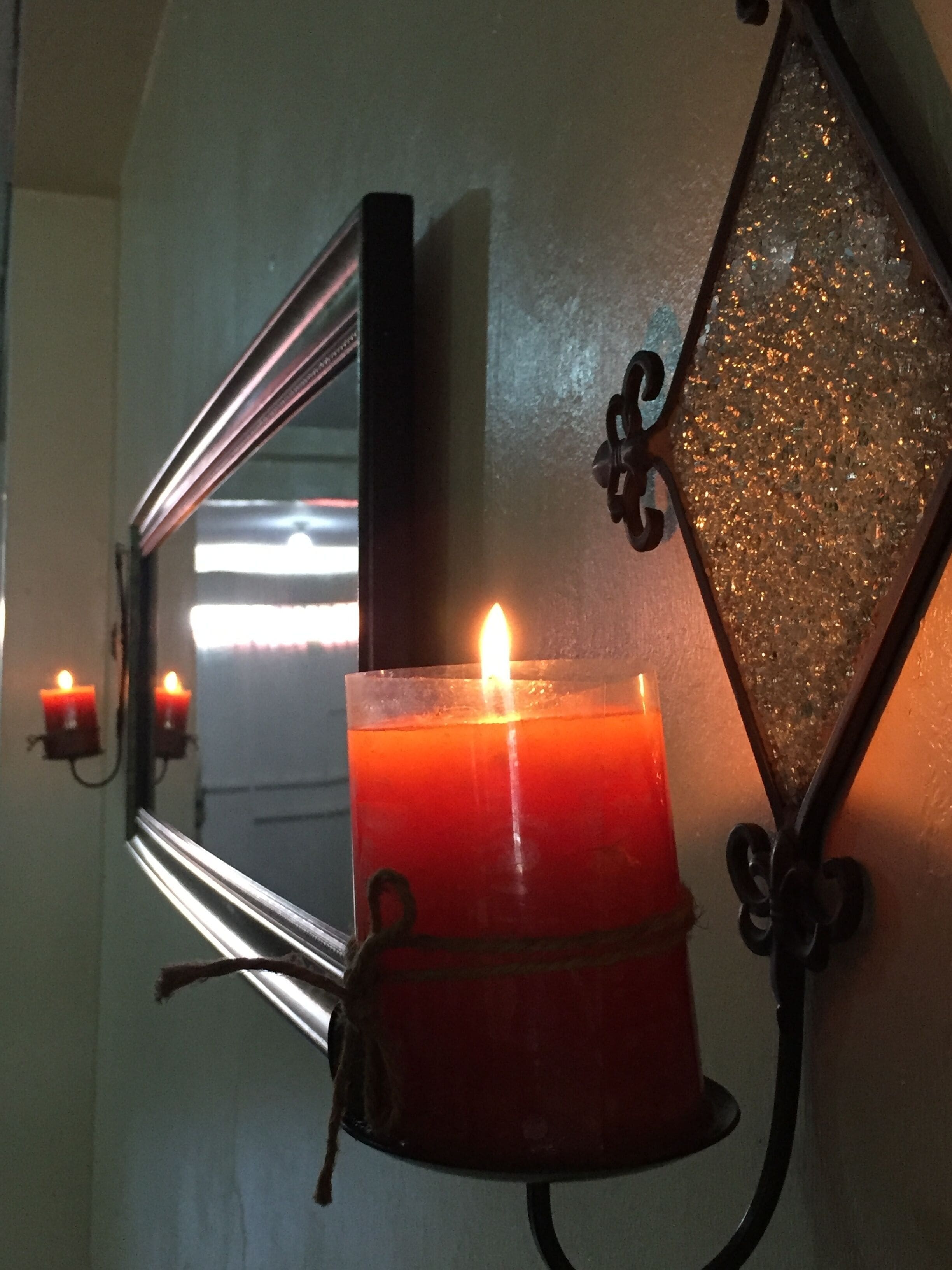 Une bougie dans un support en verre, fixée au mur, avec un miroir et un élément décoratif en métal.