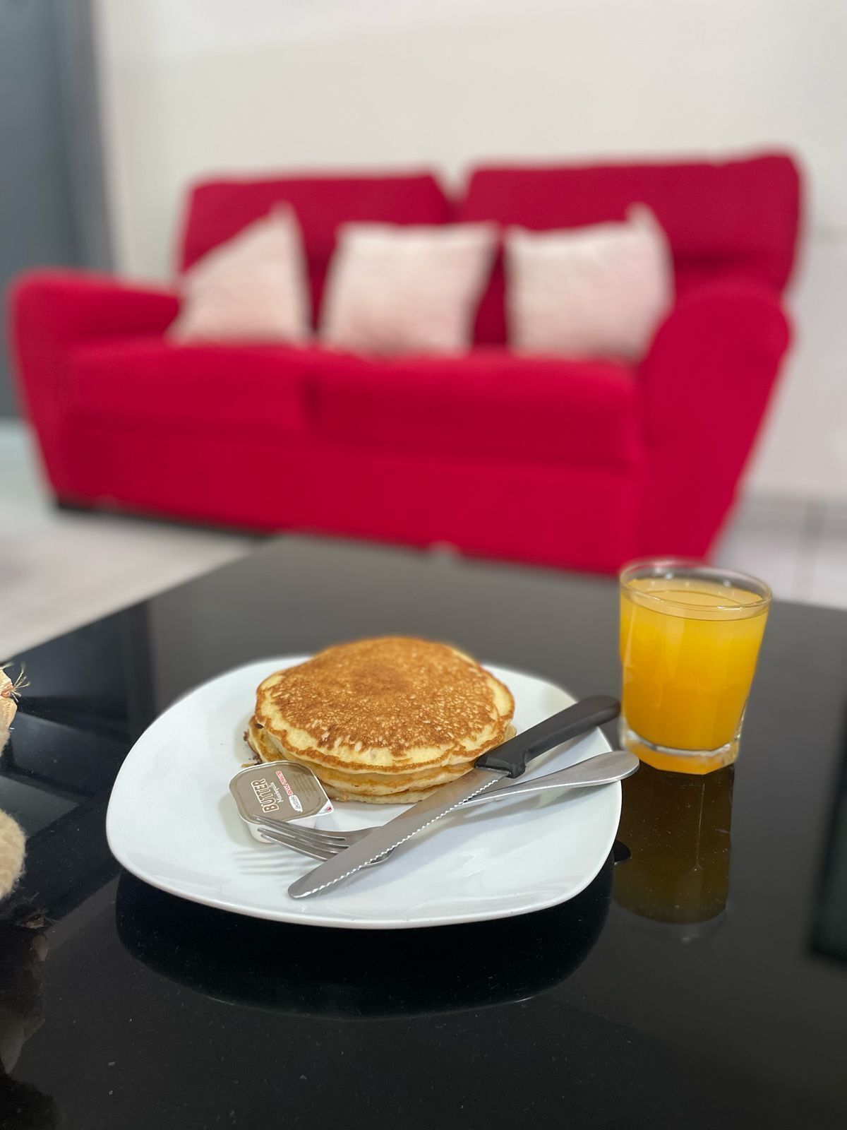 Un desayuno con tortitas en un plato, un vaso de zumo de naranja y cubiertos sobre una mesa negra.