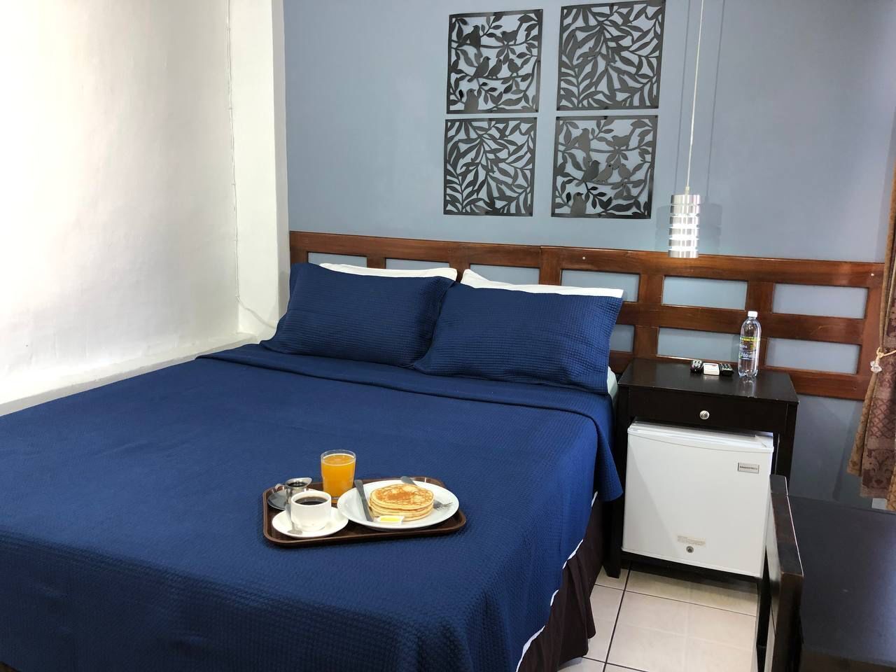 Un lit bien fait, recouvert d’une courtepointe bleue, une tablette avec un petit-déjeuner comprenant des crêpes, du café et du jus d’orange, et un petit réfrigérateur blanc posé sur une table de chevet.